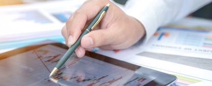 Image of a person's hand with a pen pointing at a chart on a tablet